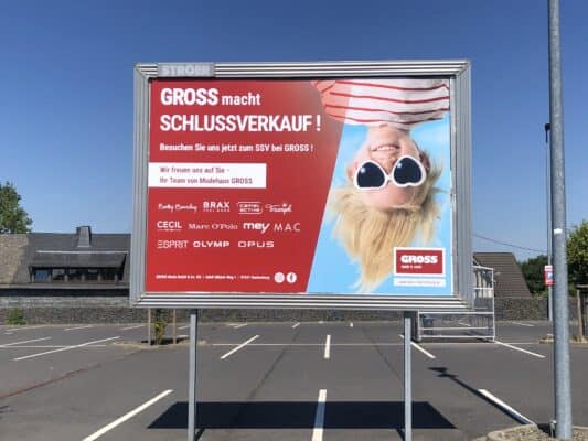 Eine große, metallene Werbetafel steht auf dem Parkplatz von groß. Auf der silbernen Tafel ist ein Werbeplakat von Gross selber befestigt, was einen Schlussverkauf ankündigt. Es ist sehr bunt und präzise gedruckt.