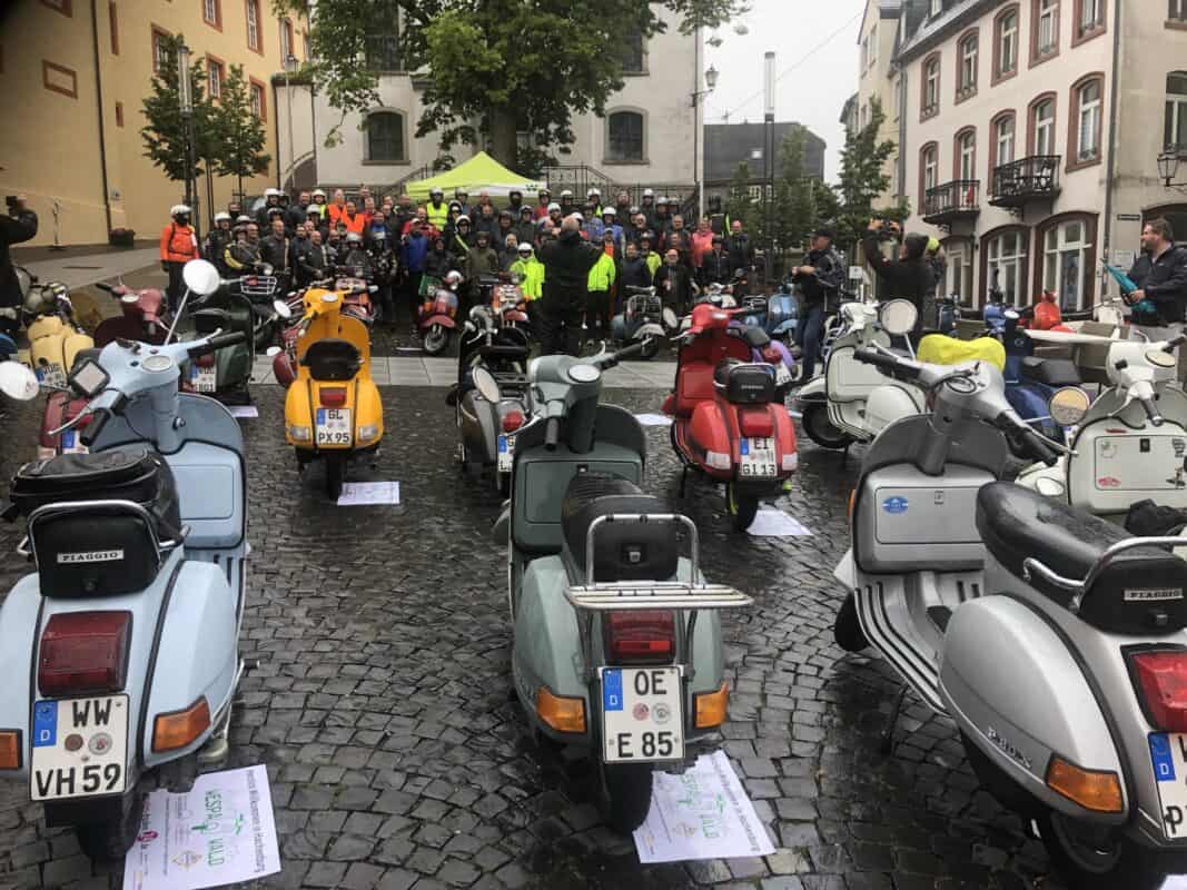 Neue und alte Vespa-Roller stehen auf dem Platz von Hachenburg vor der Kirche. Verschiedenste Modelle mit unterschiedlichen Formen, Farben und Besitzern stehen aufgereiht auf dem Pflaster mit Blickrichtung zur Kirche und verschönern den Ort. Vor der Kirche stehen deren Besitzer in dicken aber bereits nassen Jacken und Helmen, so wie vereinzelte Maschinen zwischen ihnen.