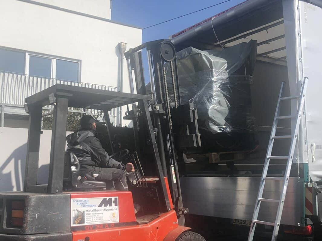 Ein roter Stapler hebt eine in dunkler Folie eingepackte Maschine aus einem LKW.