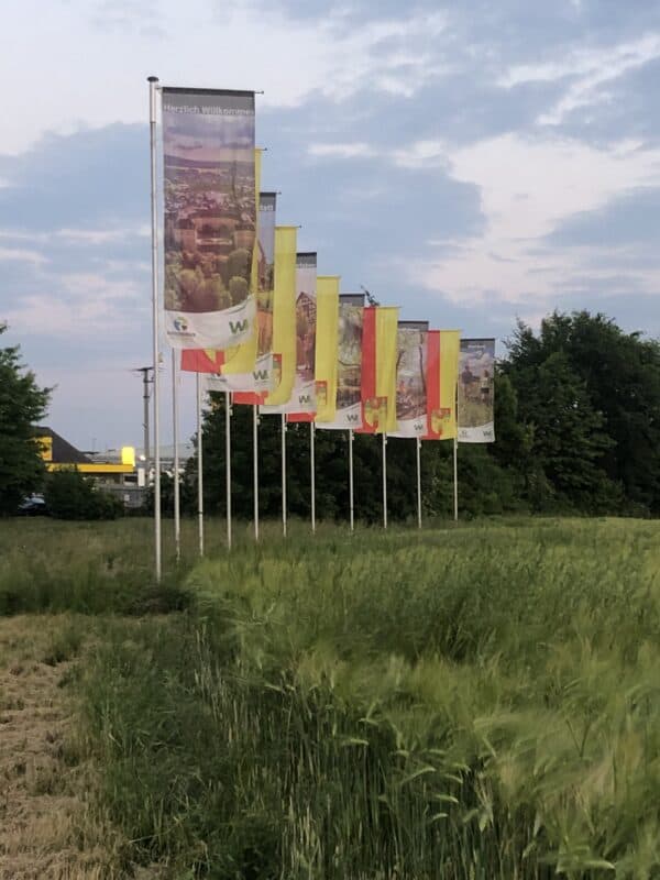 11 Fahnen stehen links geneigt in einem leichten Bogen und wehen im wind. Abwechselnd gibt es ein Bildmotiv und das gelb-rote Wappen der Stadt Hachenburg.