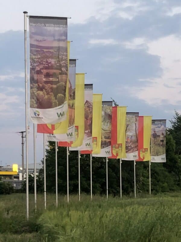 11 Fahnen stehen links geneigt in einem leichten Bogen und wehen im wind. Abwechselnd gibt es ein Bildmotiv und das gelb-rote Wappen der Stadt Hachenburg.