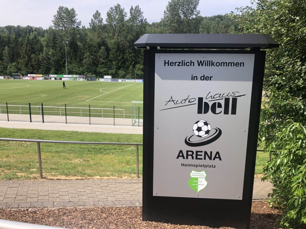 Ein dunkles Schild mit hellgrauer Platte steht vor einem Sportplatz. Drauf steht in schwarzer Schrift:"Herzlich Willkommen in der Autohaus bell Arena". Dekoriert ist es mit einem Fußball und einem grünen Wappen.