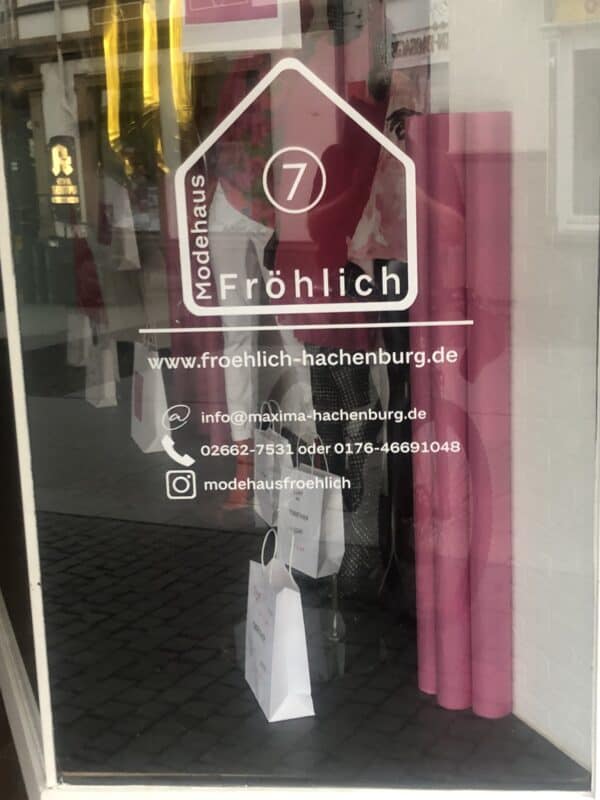 Ein Schaufensterdruck in weiß von Modehaus Fröhlich ist zu sehen. Das Logo ist ein weiß gedruckter Hausumriss mit einer eingekreisten 7 in der Mitte. Abgetrennt durch einen Strich steht unten drunter die Website, sowie eine E-Mailaddresse, Telefonnummer und ein Instagrammprofil.