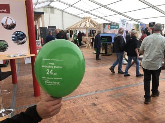 Ein hell-grüner Luftballon mit Infos zu dem Shop "wertmarken-drucken 24.de" ist zu sehen. Unter der weißen Aufschrift ist außerdem das Logo von Krüger Medienhaus, ebenfalls in weiß. Im Hintergrund befindet sich das belebte Messetreiben der Westerwälder Holztage 2022 im Sägewerk Van Roje.