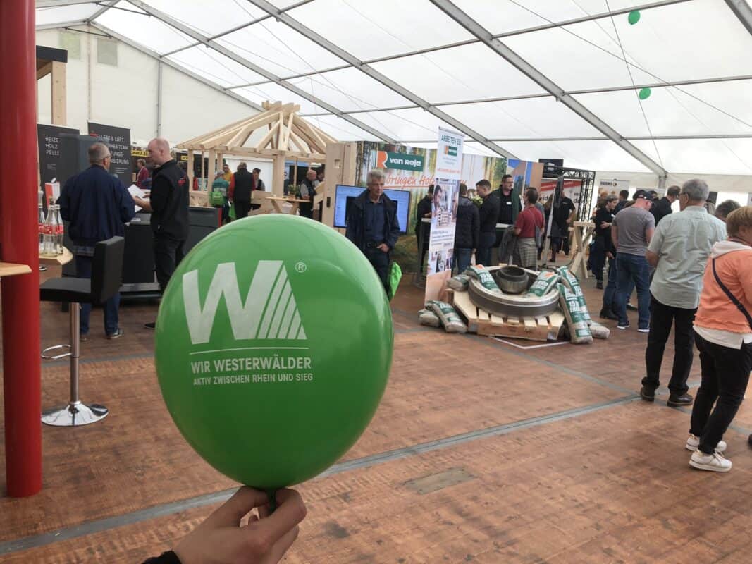 Eine gefüllte Messe ist im Hintergrund zu sehen, während im Vordergrund ein grüner Ballon steht, mit dem weißen Logo der Wir Westerwälder.