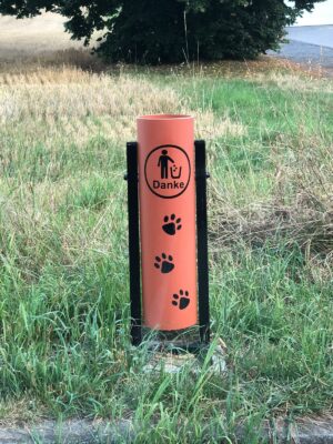 Ein orangefarbener, zylinderförmiger Mülleimer steht bei einer Wiese. Darauf gedruckt ist ein eingekreister Mensch der Müll in einen Mülleimer wirft, sowie Pfoten abdrücke darunter, beides in schwarz.